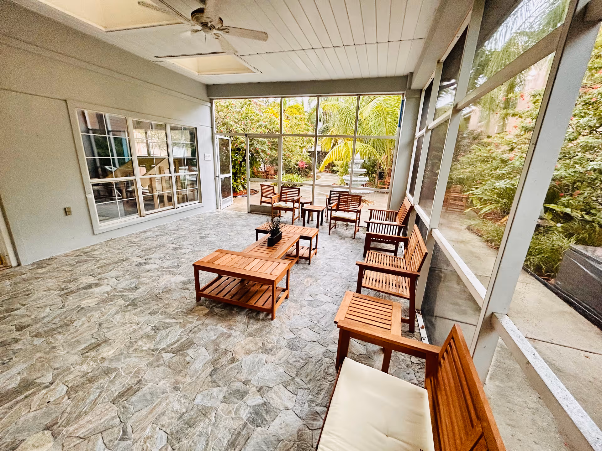 A bright and spacious screened-in patio area with stone tile flooring, wooden chairs, benches, and tables arranged for seating. The patio overlooks a garden with lush greenery, palm trees, and a water fountain in the background. A ceiling fan is mounted on the white paneled ceiling.