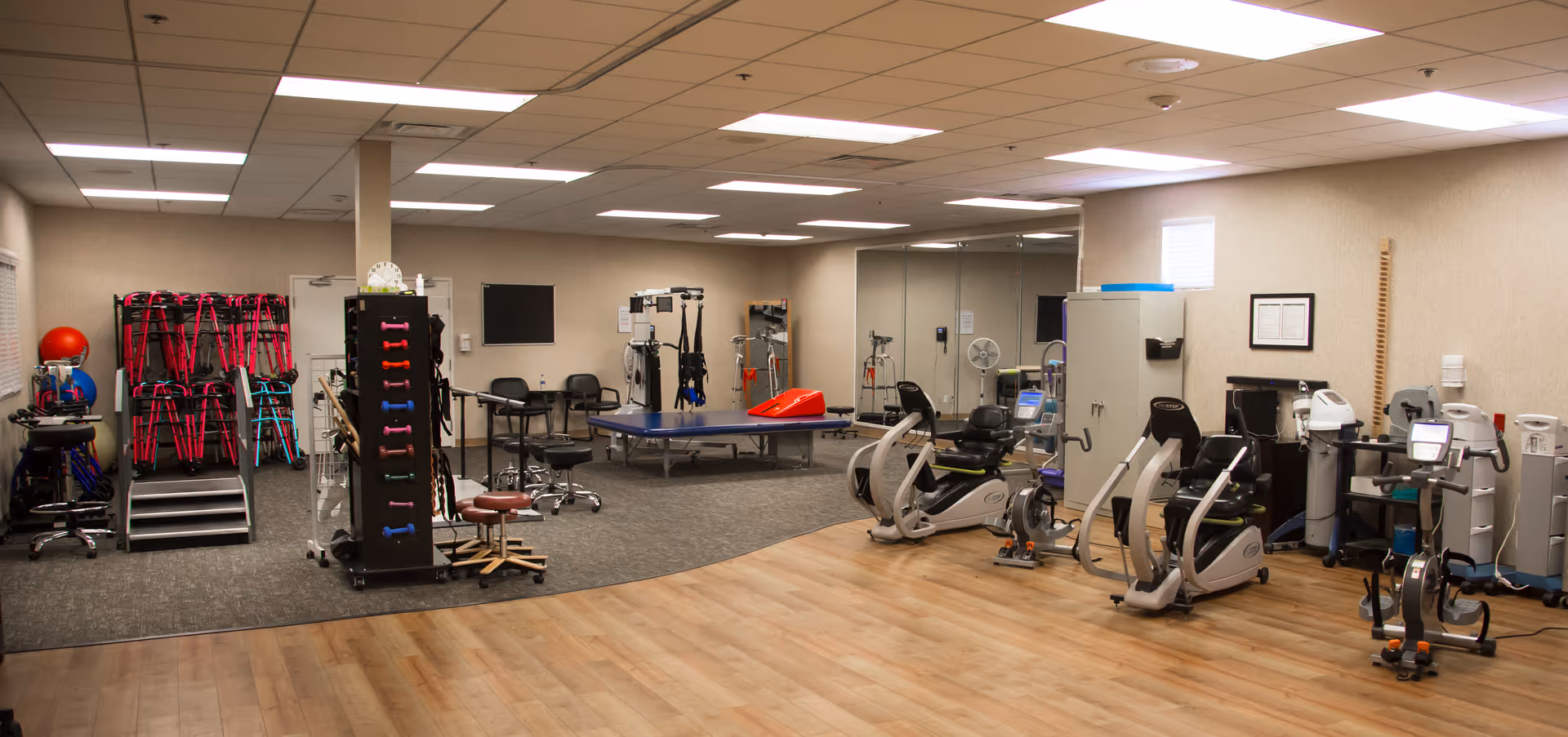 A spacious interior therapy and exercise room with cardio machines, therapy tables, weights, and stacked chairs under fluorescent lighting.