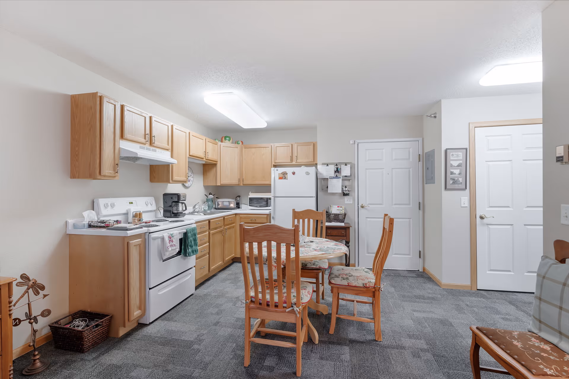 A kitchen and dining area in a senior living facility with wooden cabinets, a white stove, refrigerator, and various kitchen appliances. A round dining table with four wooden chairs featuring floral cushions is placed in the center. The room has gray carpet flooring and white walls with two white doors visible in the background.