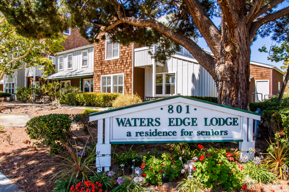 Exterior view of Waters Edge Lodge, a senior living residence, showing a white and green sign with the address 801 surrounded by plants and flowers, with the building and trees in the background.