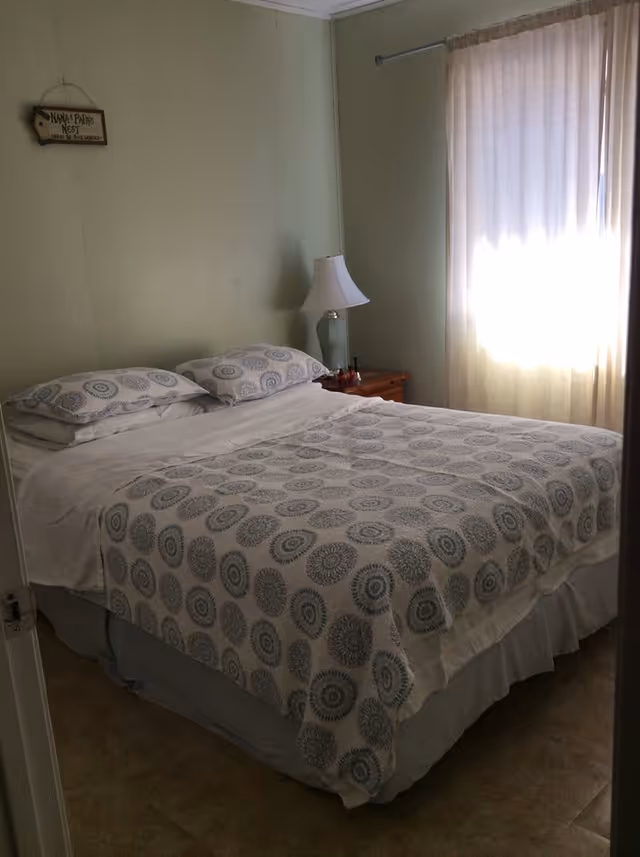 A bedroom with a neatly made bed featuring a white and blue patterned bedspread and two matching pillows. There is a wooden nightstand with a glass lamp and some small items next to the bed. A window with light-colored curtains allows sunlight to softly illuminate the room. A small decorative sign hangs on the wall above the bed.