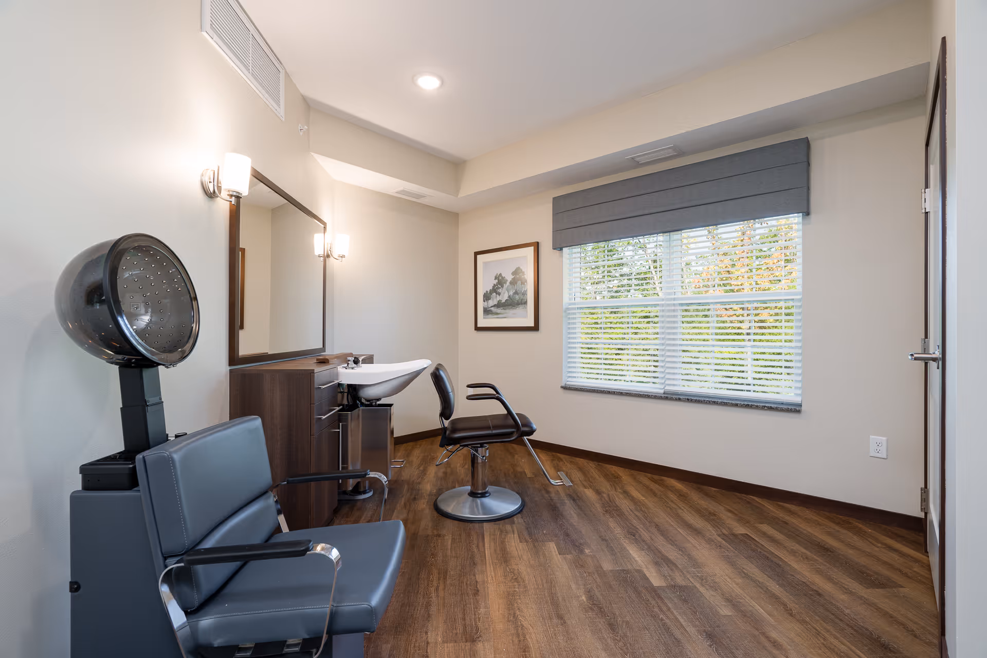 A small salon room with a hair dryer chair, a styling chair in front of a sink and mirror, wooden flooring, a window with blinds and a blue valance, and a framed picture on the wall.