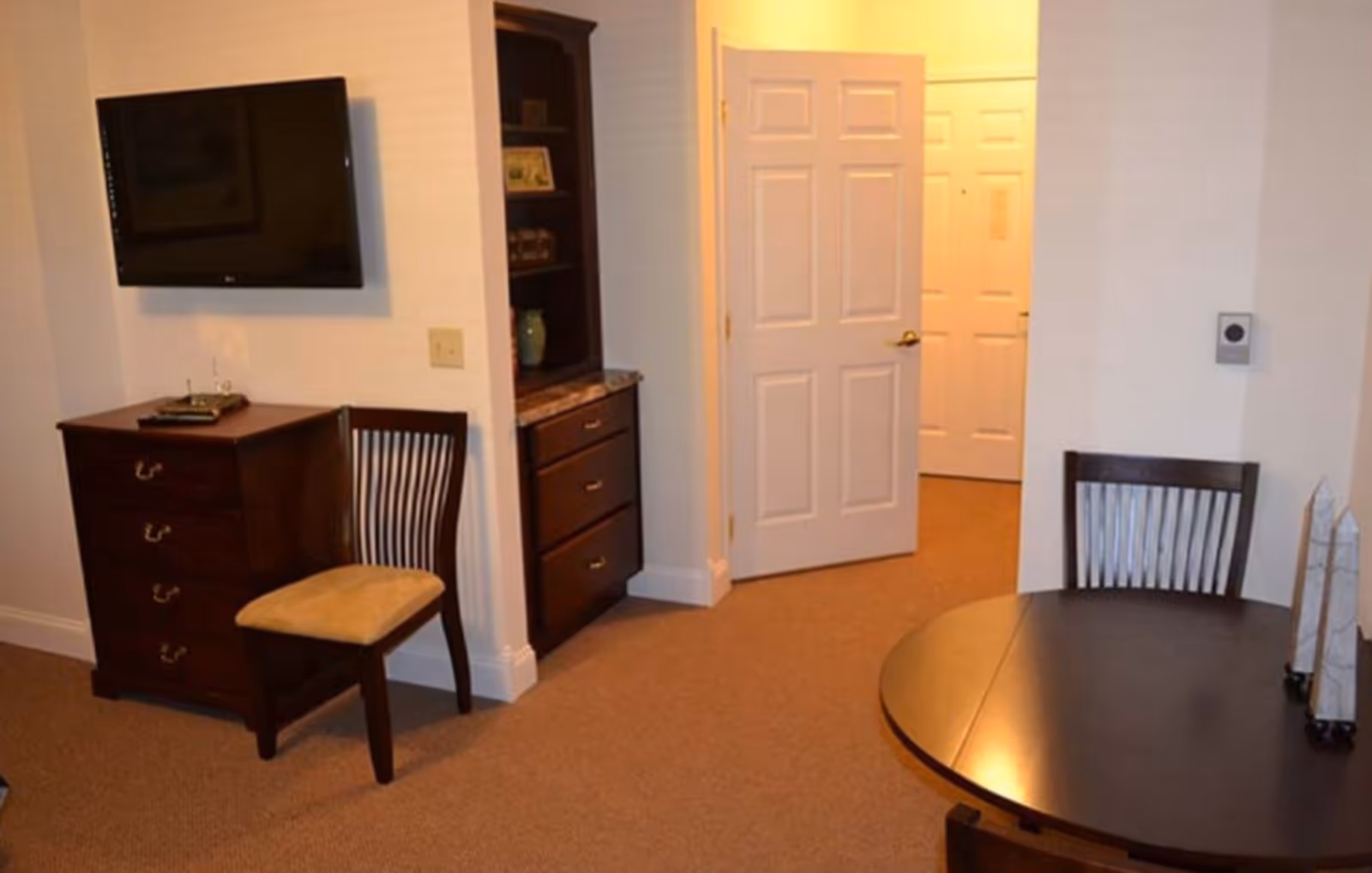 Furnished living/dining area with a wall-mounted TV, wooden chairs and dresser, a round table, and an open doorway to a hallway.