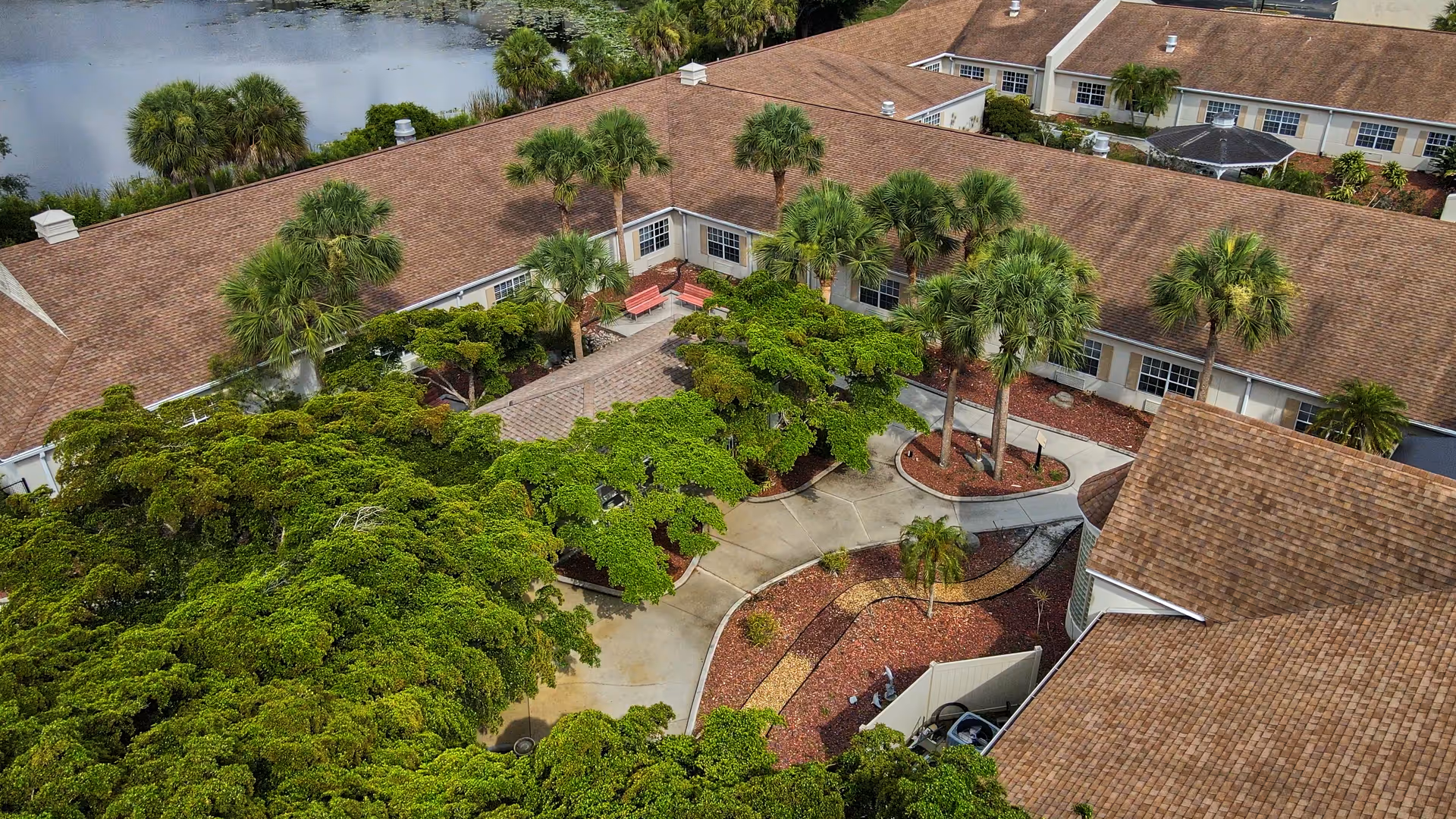 Aerial view of a senior living facility courtyard with pathways, palm trees, benches, and landscaped garden beds surrounded by single-story buildings with brown shingle roofs. A pond is visible in the background.