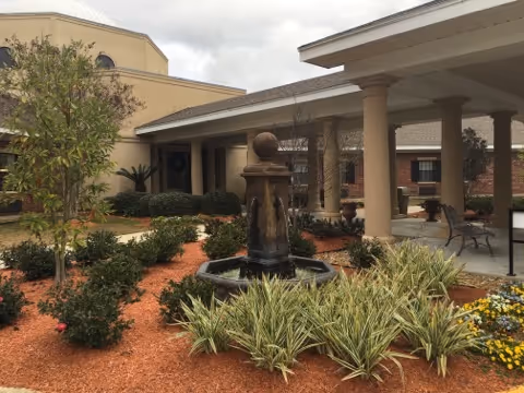 Outdoor courtyard area of Heritage Manor Opelousas featuring a central water fountain surrounded by landscaped plants and mulch, with a covered patio supported by columns and seating benches.