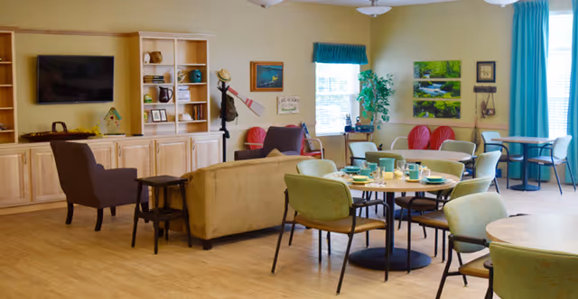 Bright communal dining and lounge area with round dining tables, chairs, sofas, a TV and shelving.