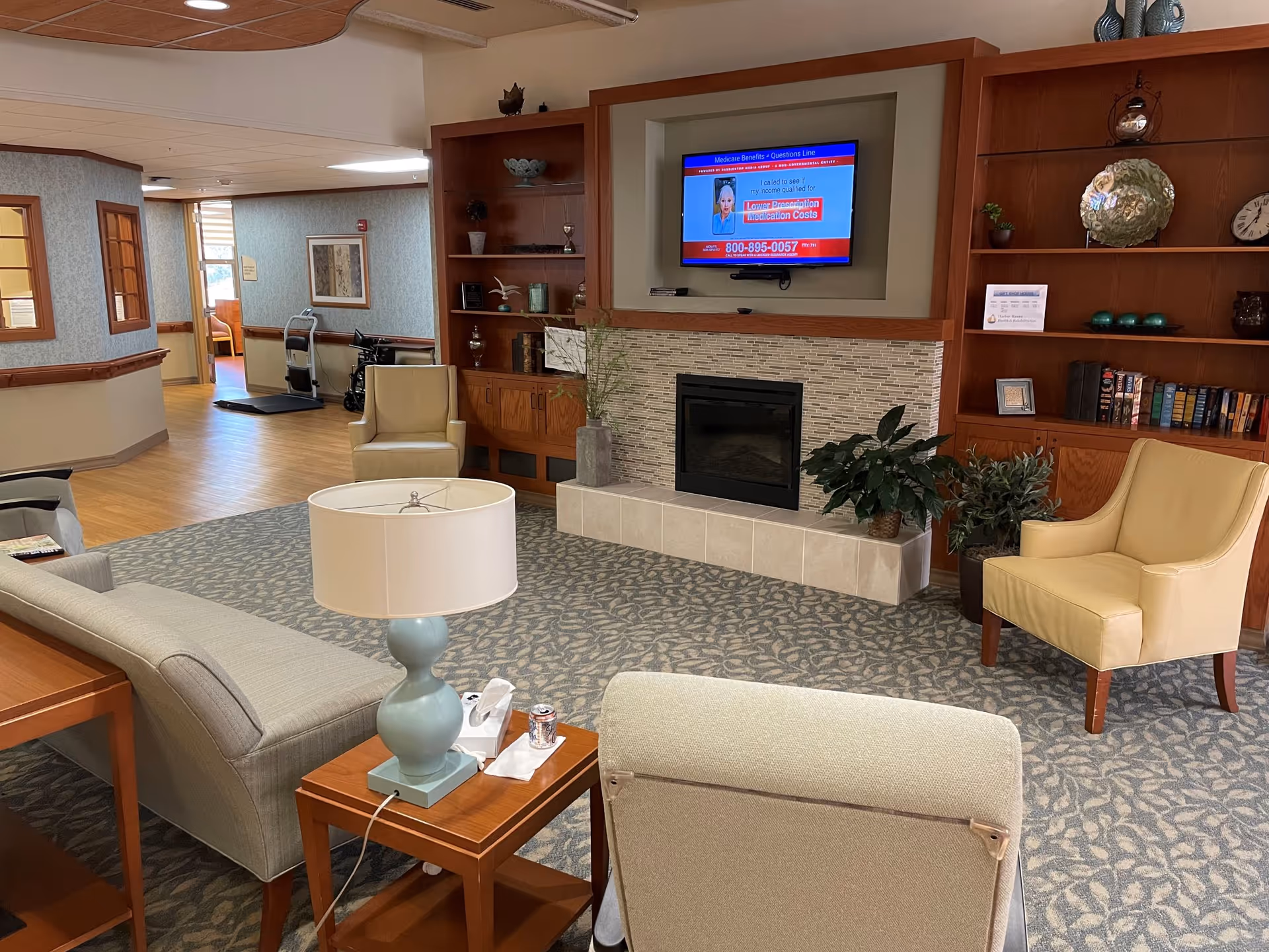A cozy living room area in a senior living facility with a fireplace and a wall-mounted TV above it. The room has a patterned carpet, a light blue sofa, beige armchairs, a wooden side table with a lamp, and built-in wooden shelves with decorative items and books. There is a hallway visible in the background with a walker and wheelchair near the entrance.