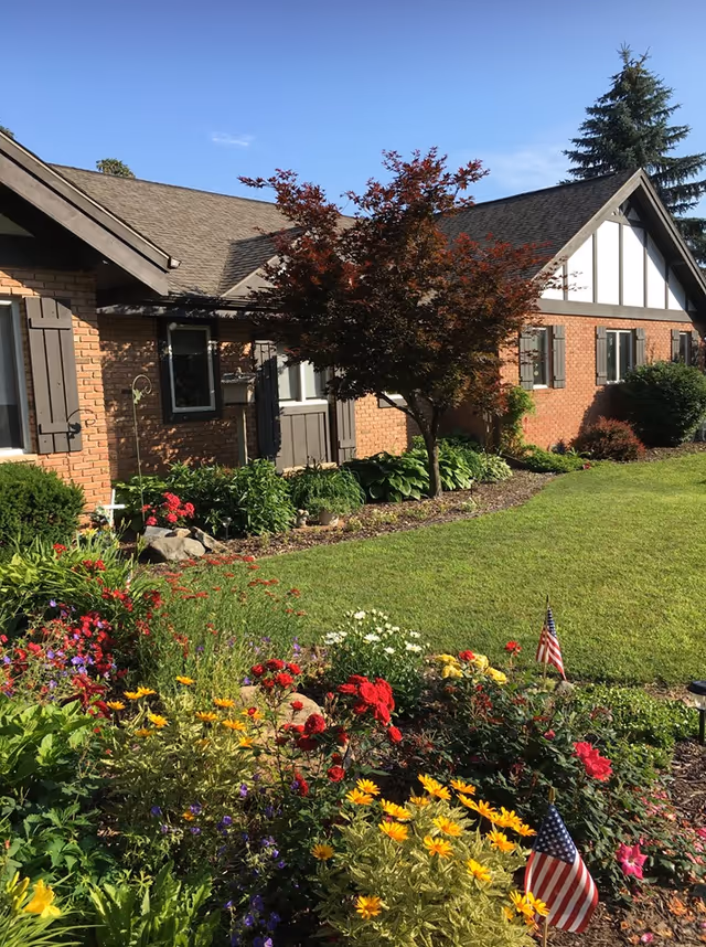 Brick single-story building with a landscaped front yard, colorful flower beds, a small tree, and two American flags.