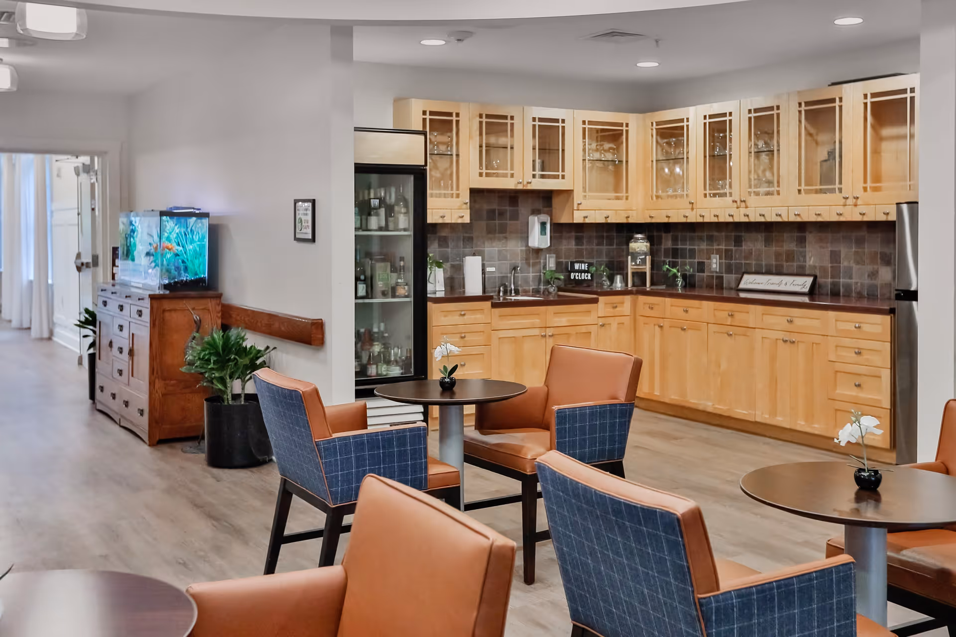 A cozy assisted living common area with a kitchenette featuring wooden cabinets and a tiled backsplash. There are several round tables with comfortable chairs upholstered in brown leather and blue fabric. A wooden sideboard with a fish tank on top is visible along the hallway, and a glass-front refrigerator stocked with beverages is integrated into the kitchenette.