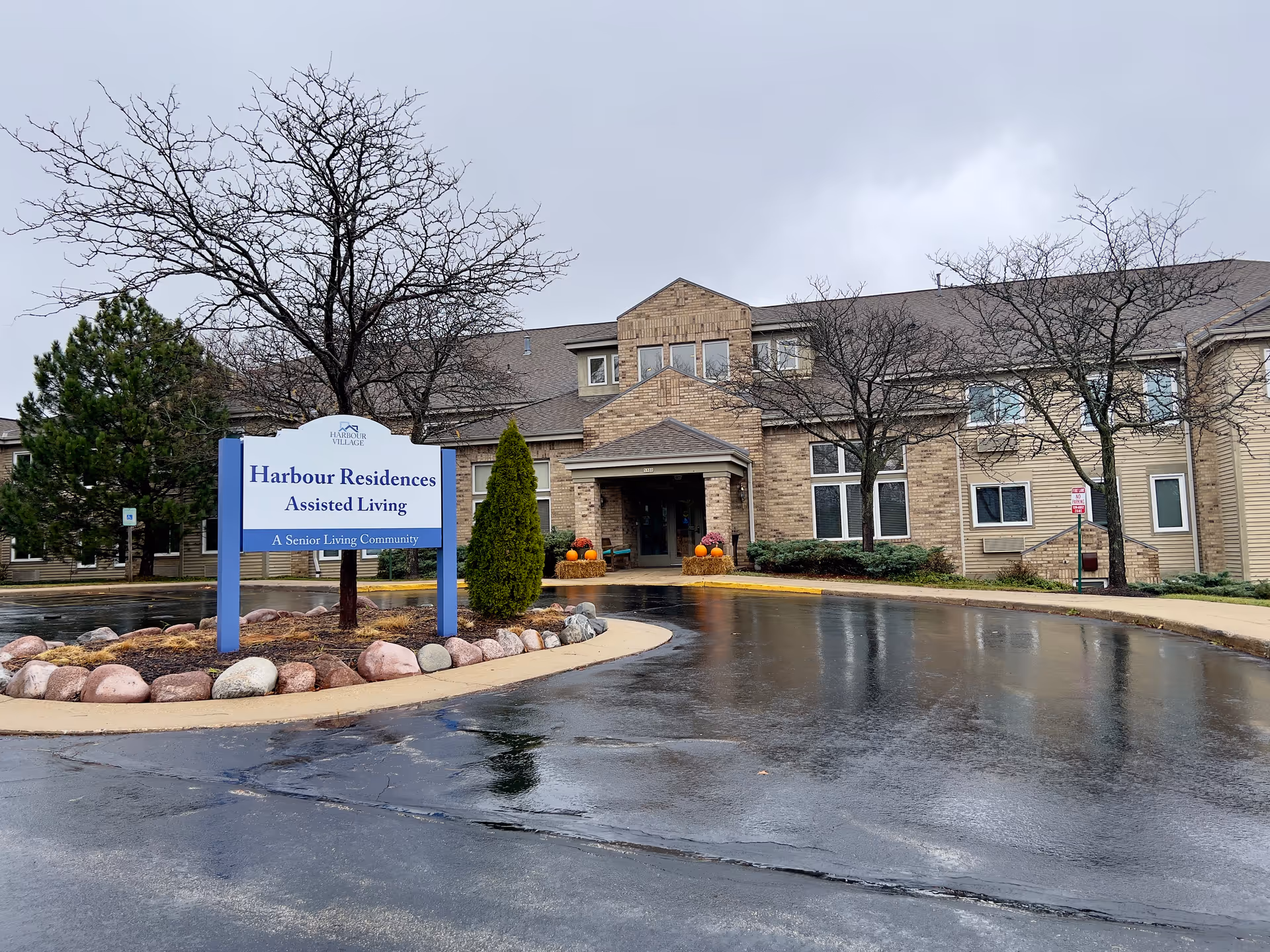 Exterior view of Harbour Village assisted living facility on a cloudy day. The building is two stories with a brick and siding facade. In front of the building is a sign that reads 'Harbour Residences Assisted Living, A Senior Living Community.' There are leafless trees and some landscaping with rocks and bushes around the sign and building entrance. The driveway is wet, reflecting the overcast sky.