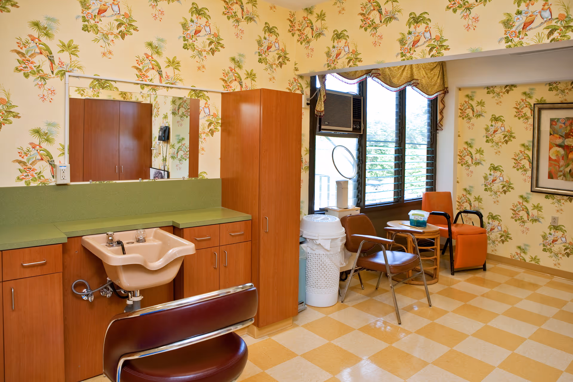 A room with floral wallpaper featuring birds and flowers, a green countertop with wooden cabinets, a beige sink with a maroon chair in front, and a large mirror above the sink. To the right, there are two chairs, a small round table, a window with an air conditioning unit, and a framed floral painting on the wall.