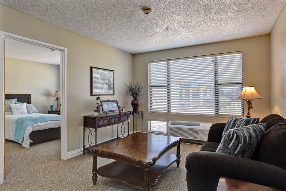 Bright living room with a dark sofa, wooden coffee table, console table and large window with blinds, and a bedroom visible through an open doorway.