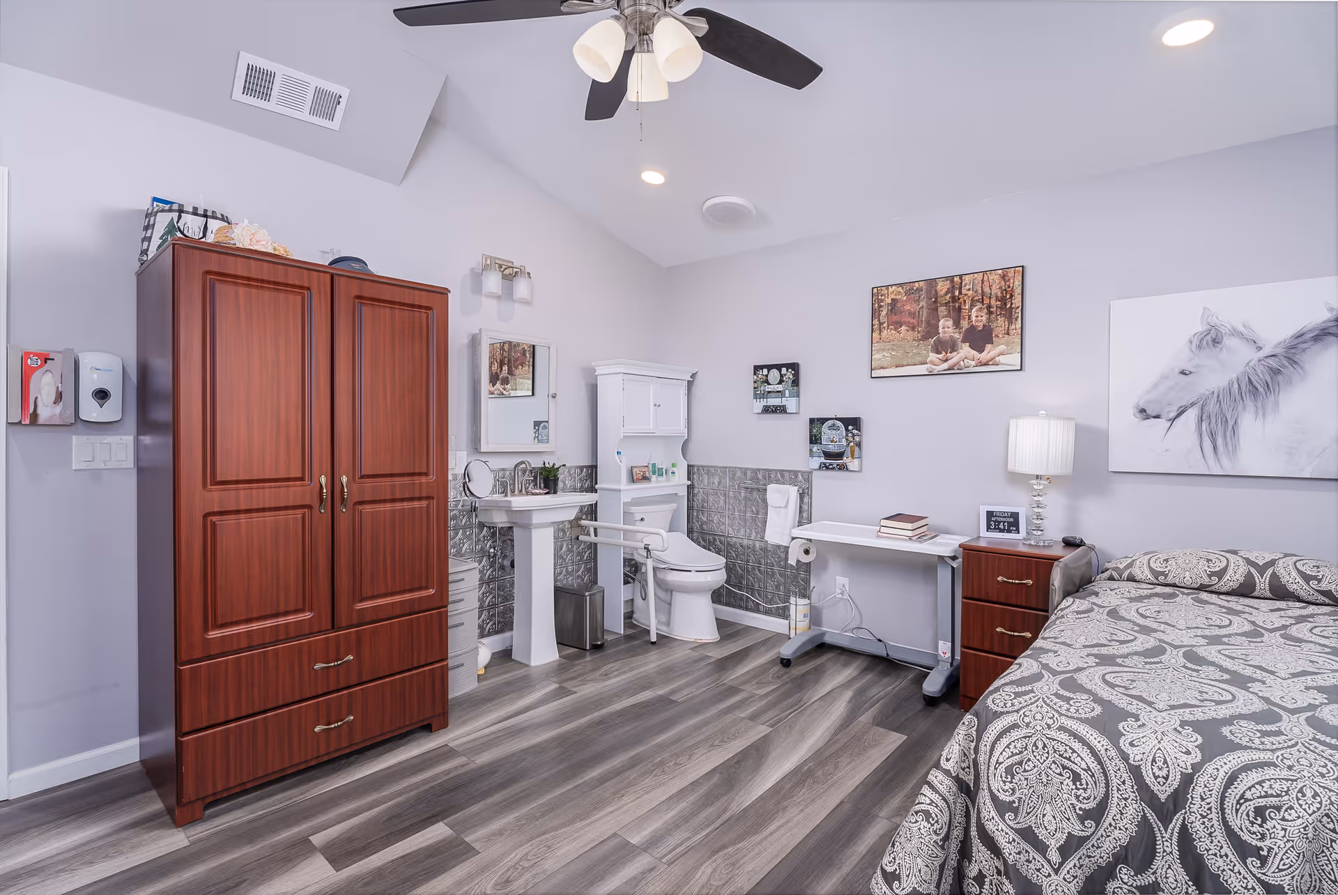 A well-lit bedroom in an elder care facility featuring a bed with patterned bedding, a wooden nightstand with a lamp and clock, a small desk with books, a white toilet and sink with a mirror above, a wooden wardrobe, and wall decorations including a photo of two children and a painting of a white horse.