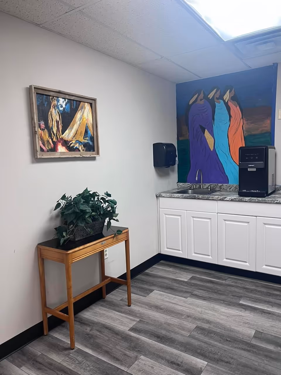 Interior corner of a room with a small wooden table holding a potted plant, white cabinets with a granite countertop, a sink, a water dispenser, a wall-mounted paper towel dispenser, and two colorful paintings on the walls.