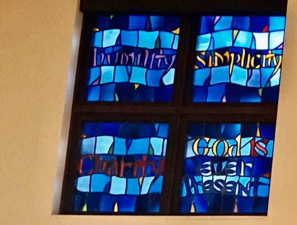 A stained glass window divided into four sections with blue and purple hues. The sections contain words in different colors: 'Humility', 'Simplicity', 'Clarity', and 'God is ever present'.