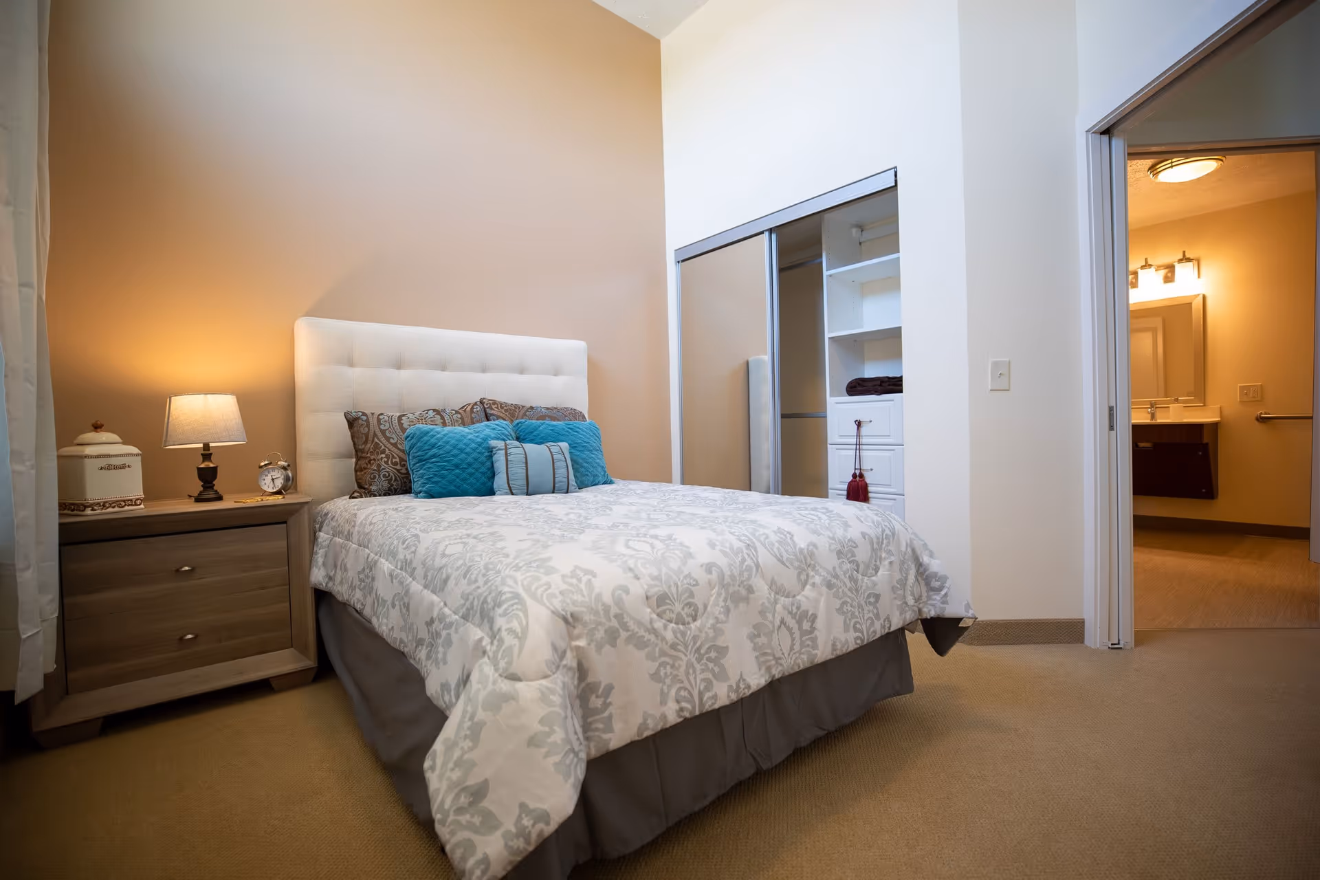 A neatly made bed with a white and gray patterned comforter and several decorative pillows in blue and brown tones. To the left of the bed is a wooden nightstand with a lamp, a small clock, and a decorative container. On the right side, there is an open closet with sliding mirrored doors and built-in shelves. An open doorway leads to a bathroom with a sink, mirror, and lighting fixture.