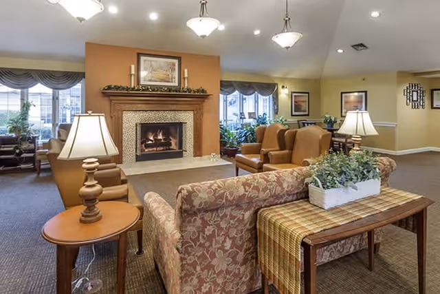 A cozy living room area in a senior living facility featuring a lit fireplace with a mantel decorated with candles and a framed picture above it. The room has multiple comfortable armchairs and sofas arranged around the fireplace, side tables with lamps, large windows with curtains, and various plants and framed artwork on the walls.