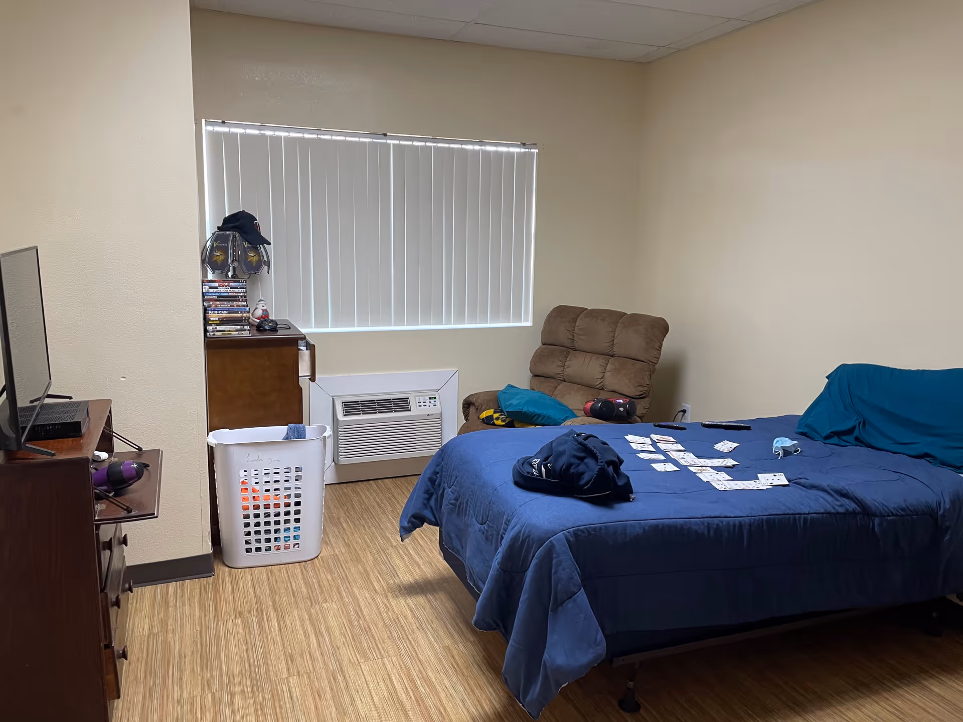 A bedroom with a bed covered in a blue comforter and a teal pillow. Playing cards are spread out on the bed along with a black cap and a face mask. There is a brown cushioned chair in the corner next to a window with vertical blinds. A wooden dresser with a stack of DVDs and a hat on top is next to a white laundry basket. A TV is placed on a wooden stand on the left side of the room. The floor has a wood-like finish and the walls are beige.