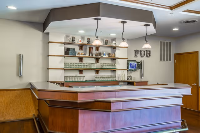Interior view of a wooden bar area with shelves holding glassware and decorative items. Three pendant lights hang above the bar counter. A wall-mounted phone and a sign spelling 'PUB' are visible on the wall. The room has a wooden door and neutral-colored walls.