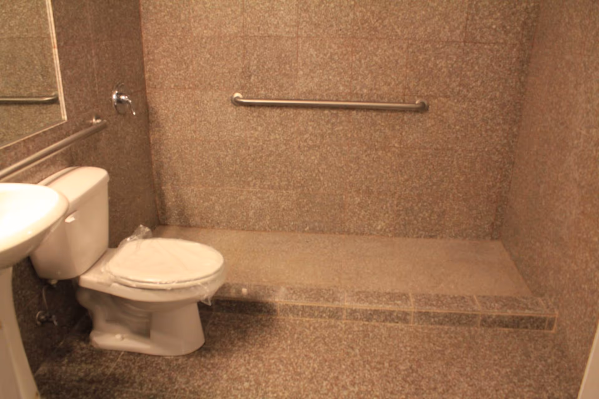 A bathroom with a white toilet and pedestal sink. The toilet seat is covered with plastic. The walls and floor are covered with brownish tiles. There is a metal grab bar on the wall next to the toilet and another grab bar on the back wall above a raised tiled platform.