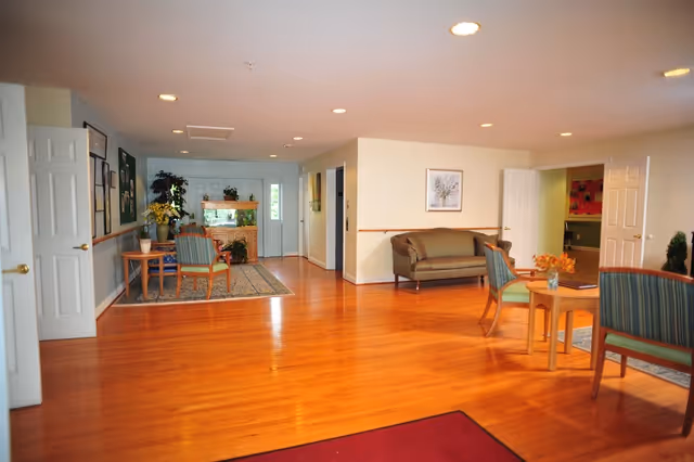 A spacious and well-lit common area with polished wooden floors, several chairs and small tables arranged for seating, a green couch against the wall, framed artwork, and potted plants. Multiple open doors lead to other rooms, and the area has recessed ceiling lights.