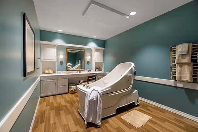 A spacious bathroom with teal walls and wood flooring featuring a large white therapeutic bathtub with a white robe hanging on its side. The room includes a vanity with a mirror, two wall sconces, and cabinets. Towels are neatly arranged on a wooden towel rack mounted on the wall.