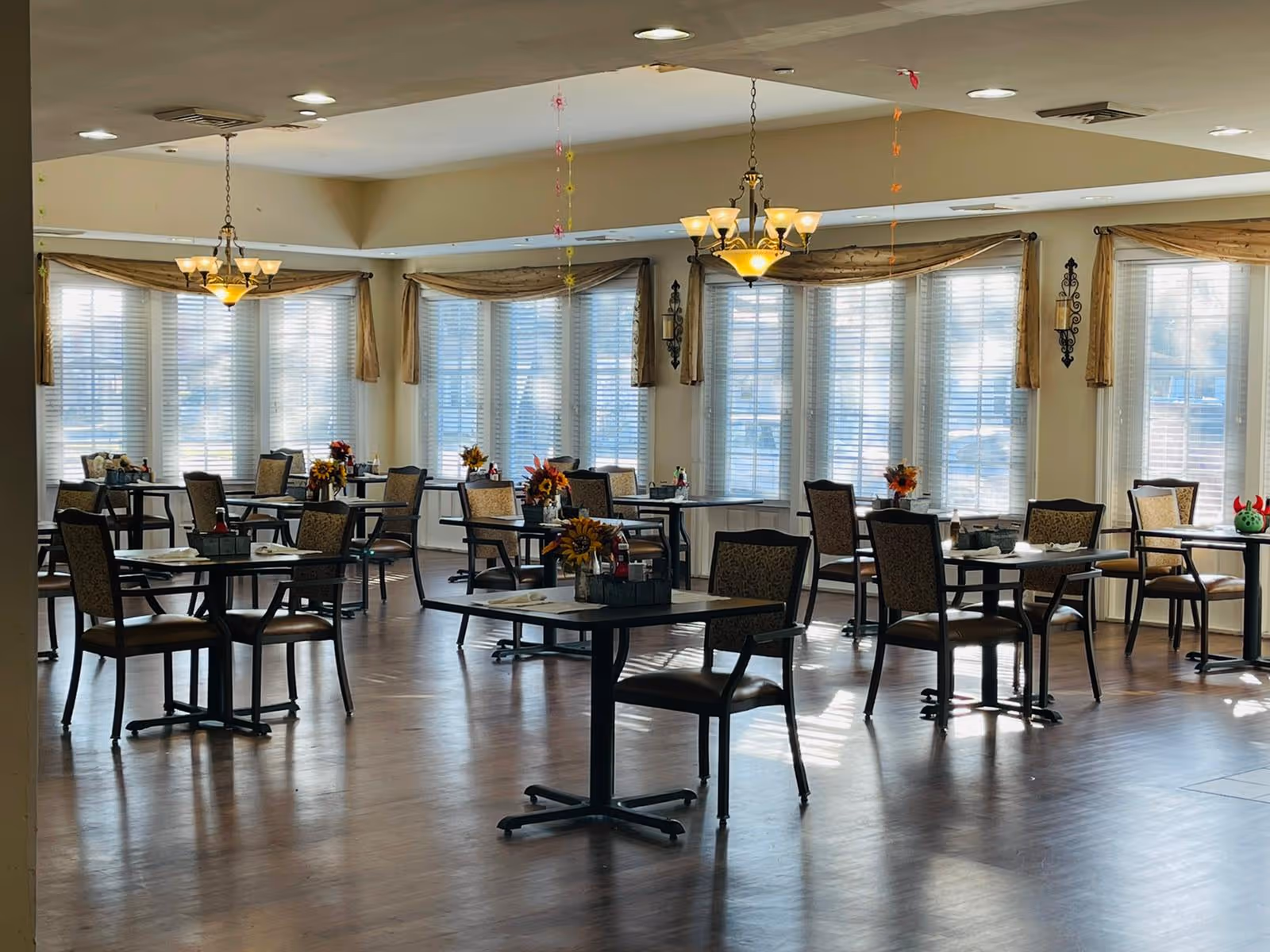 A bright dining room with multiple tables and chairs arranged neatly. Each table has a small floral centerpiece. Large windows with blinds and valances allow natural light to fill the room. Two chandeliers hang from the ceiling providing additional lighting.
