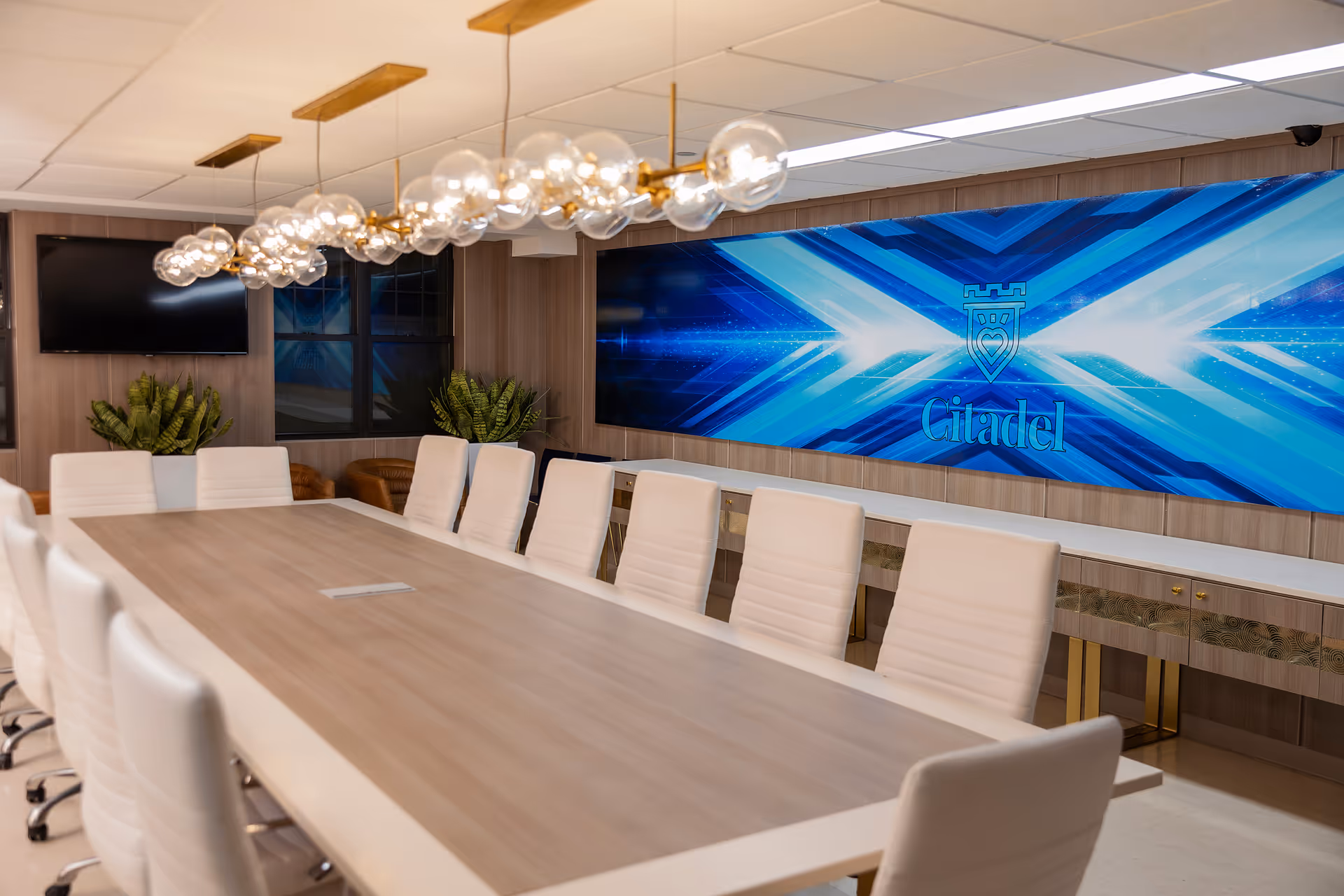 Modern conference room with a long wood table, white chairs, pendant lights, plants, and a large blue digital screen displaying the Citadel logo.