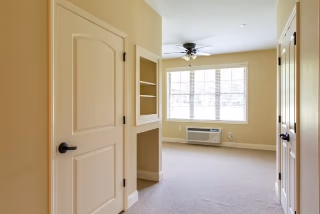 Empty room with beige walls and carpet, featuring two closed doors on the left and right sides, a built-in shelving niche, a large window with blinds, a ceiling fan with light, and a wall-mounted air conditioning unit below the window.