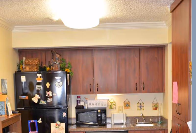 A small kitchen area with dark wooden cabinets, a black refrigerator decorated with magnets, a microwave, a toaster, and a sink. There are decorative items on top of the refrigerator and small hanging decorations on the wall above the countertop.