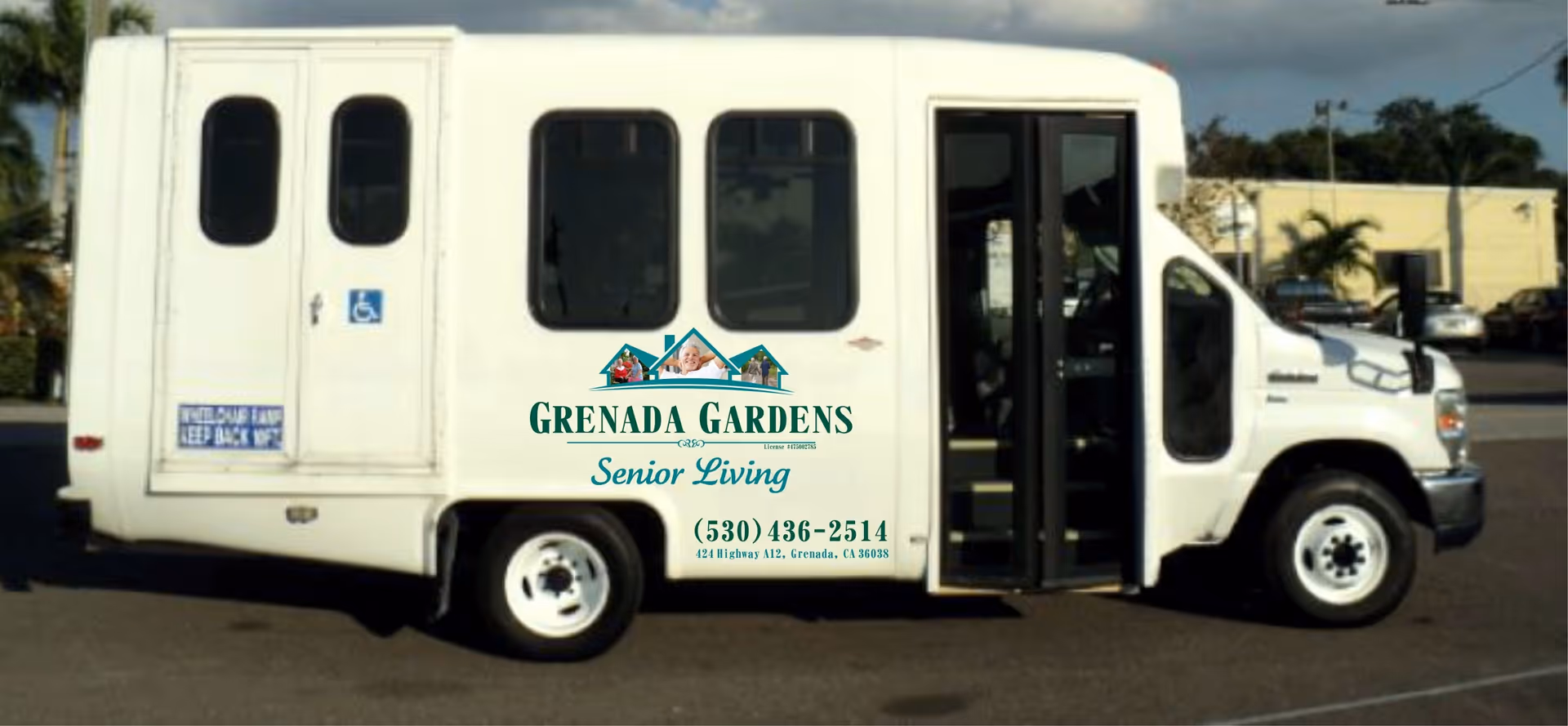 White shuttle bus parked outdoors with the logo and contact information for Grenada Gardens Senior Living on its side. The bus has a wheelchair accessibility sign near the rear door and an open front passenger door.
