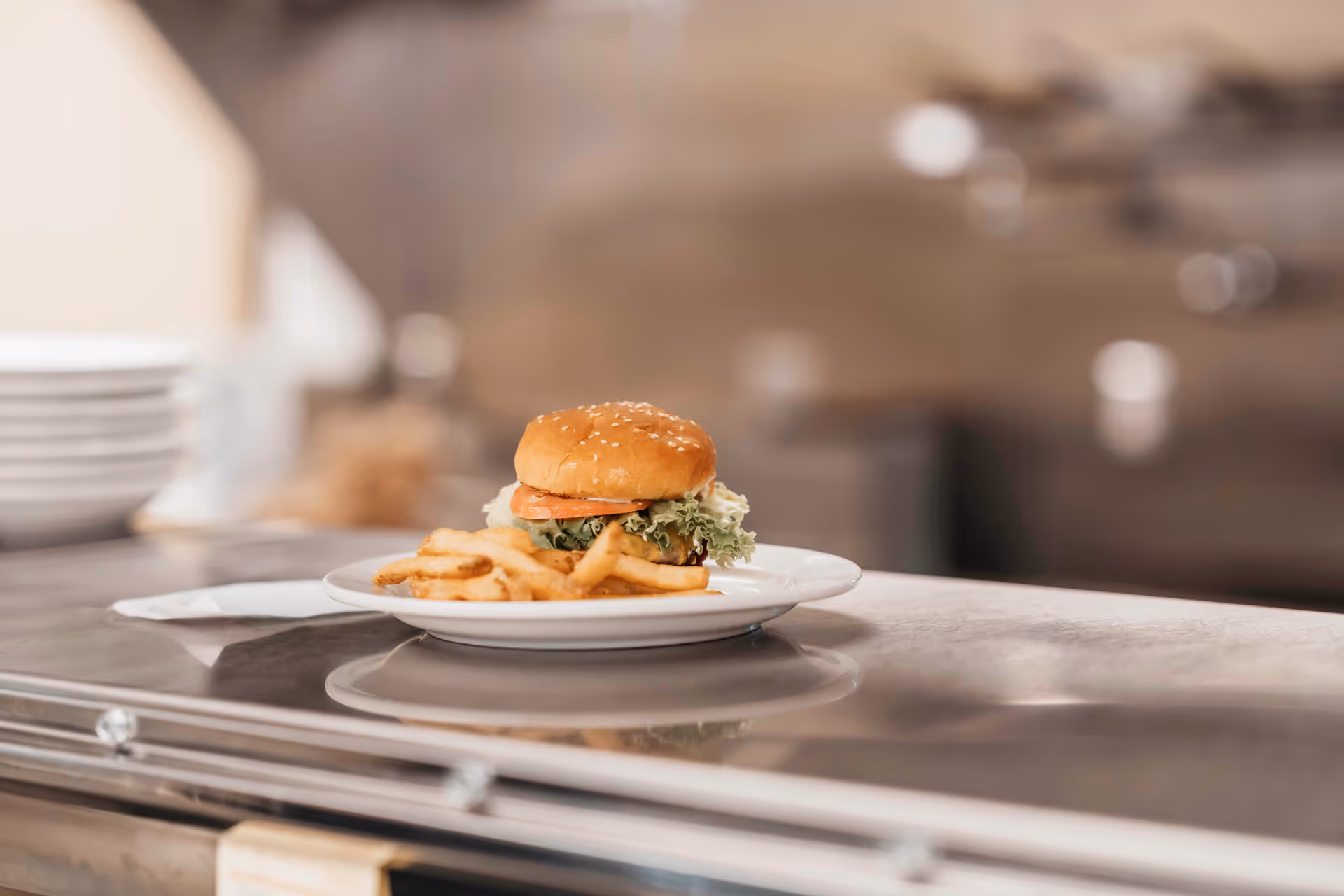 A plate with a hamburger and French fries placed on a stainless steel counter in a kitchen setting. The hamburger has a sesame seed bun, lettuce, tomato, and cheese. In the background, there are stacked white plates and blurred kitchen equipment.