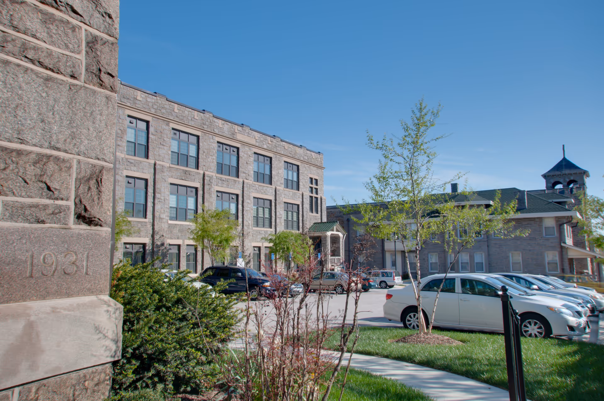 Exterior view of the John Manley House senior living facility showing a multi-story stone building with numerous windows, a parking lot with several cars, green trees, and a clear blue sky.