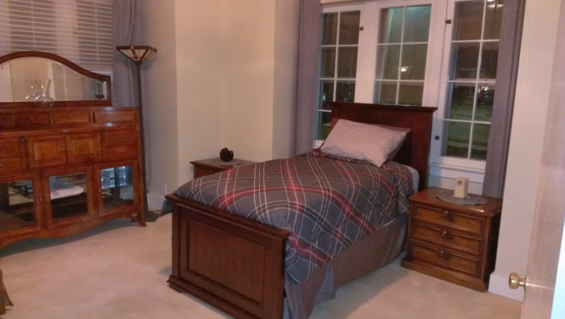 A bedroom with a single wooden bed covered with a plaid comforter and a pillow. There are two wooden nightstands on either side of the bed, one with a candle and the other with a small decorative item. A wooden dresser with a mirror is positioned against the wall, and a tall floor lamp stands in the corner. The room has large windows with curtains and light-colored walls and carpet.