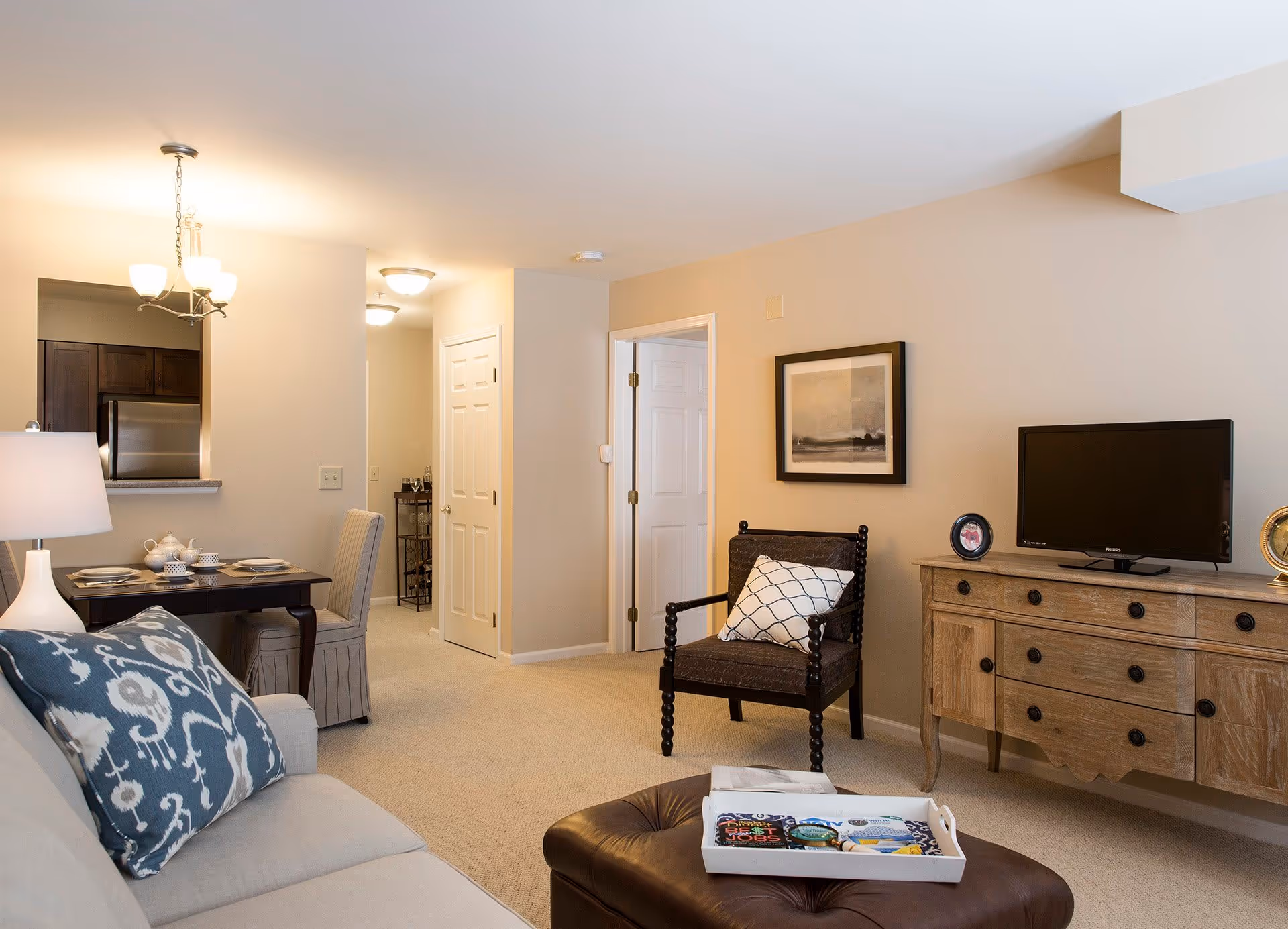 A cozy living room area in a senior living facility with a beige sofa adorned with patterned pillows, a dark wooden chair with a white cushion, a wooden sideboard with a small TV and framed photos, and a brown ottoman with a tray holding magazines. In the background, there is a small dining table set for two and a view into a kitchen area with stainless steel appliances.
