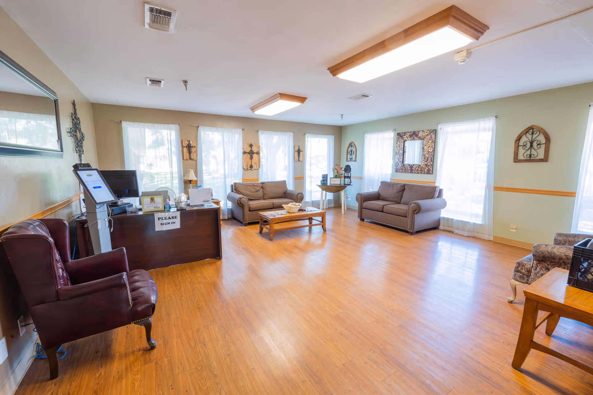 A bright and spacious common area in a senior living facility with wooden flooring, multiple large windows with sheer white curtains, two brown sofas, a coffee table, a desk with a sign-in kiosk, and various decorative wall hangings and mirrors.