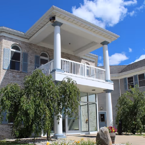 Front entrance of a two-story senior living building with white columns, a covered balcony, glass doors, and landscaping.