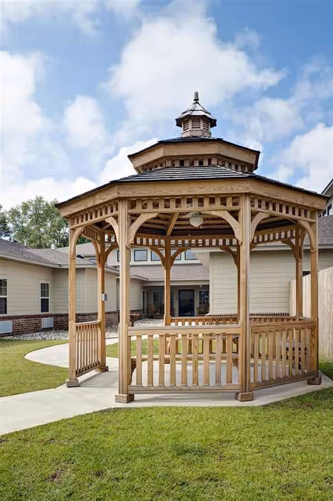 Wooden gazebo in a grassy courtyard with a walkway and residential building behind.