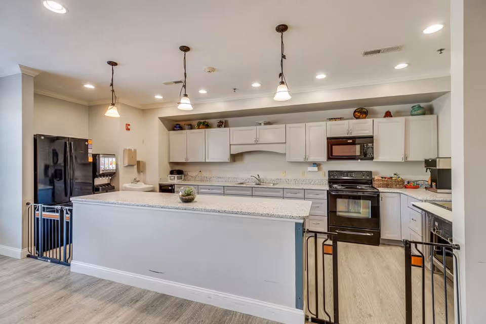 A modern kitchen area in an assisted living facility with white cabinets, a black refrigerator, black stove, microwave, and dishwasher. There is a large island countertop in the center with a small plant on it. Three pendant lights hang from the ceiling above the island. The floor is light-colored wood, and there are safety gates at the entrance to the kitchen.