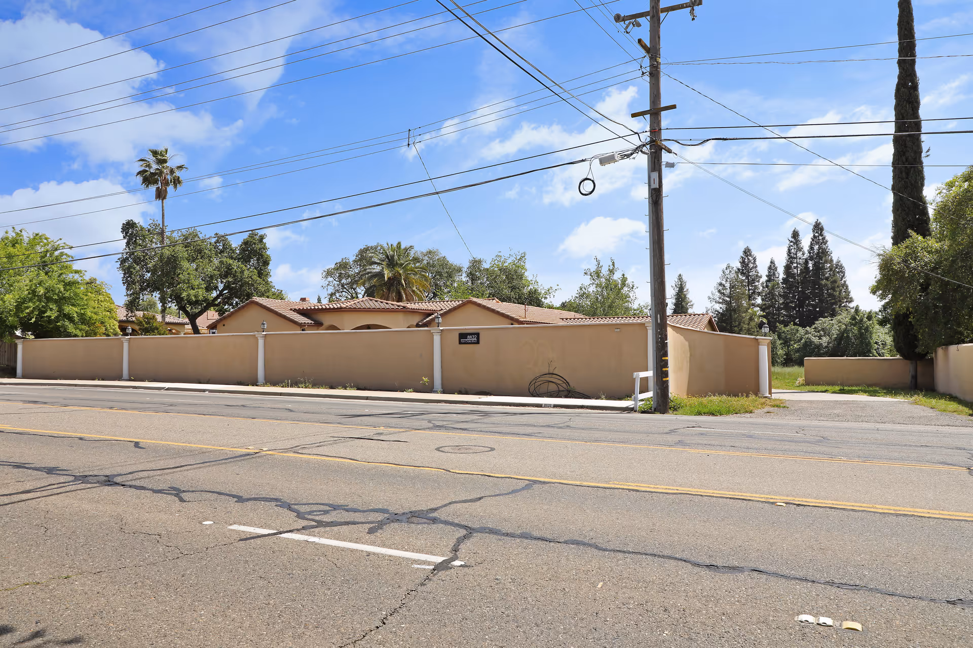 Street view of a single-story building behind a stucco wall with a tiled roof, trees, and utility poles under a blue sky.