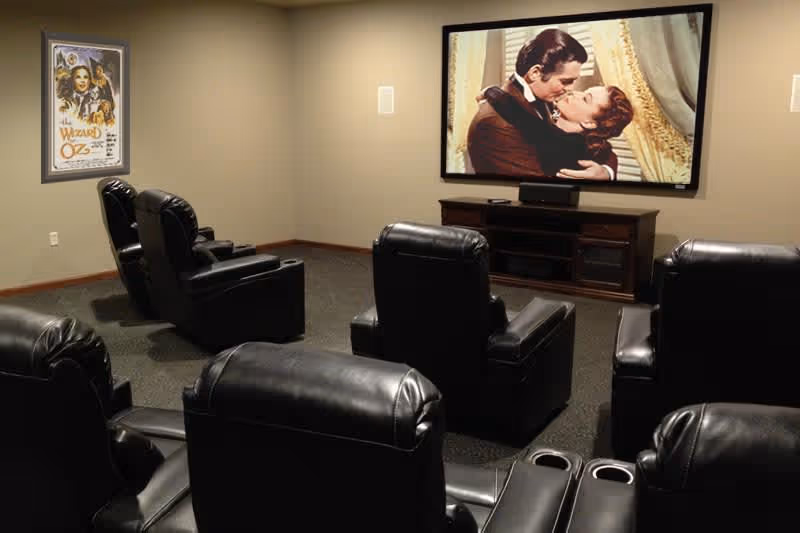 A small theater room with six black leather recliner chairs arranged in two rows facing a large screen showing a romantic movie scene. On the left wall, there is a framed poster of The Wizard of Oz.