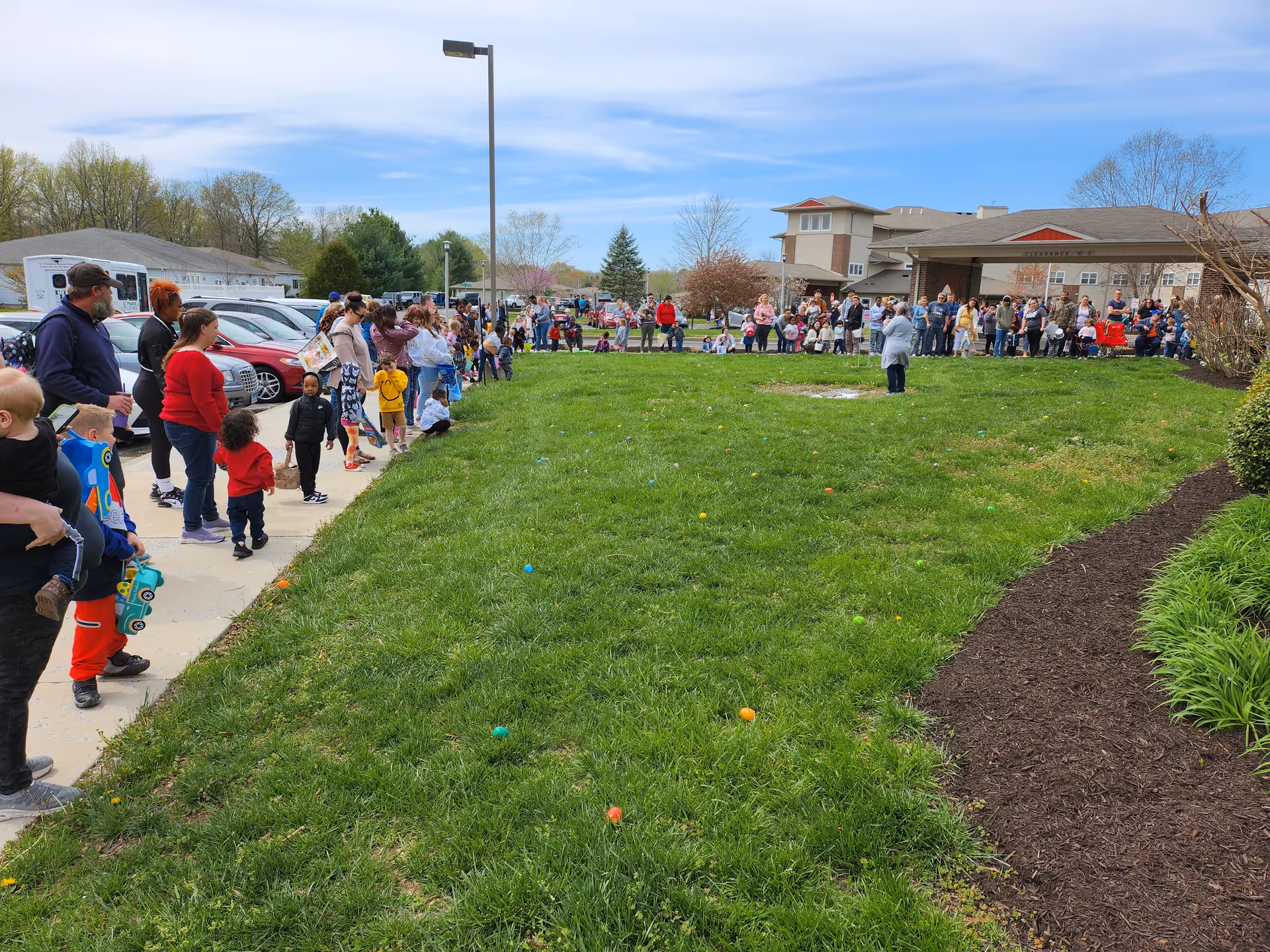 A large crowd of people and children gathered along a sidewalk and grassy lawn outside a senior living building for an outdoor event.