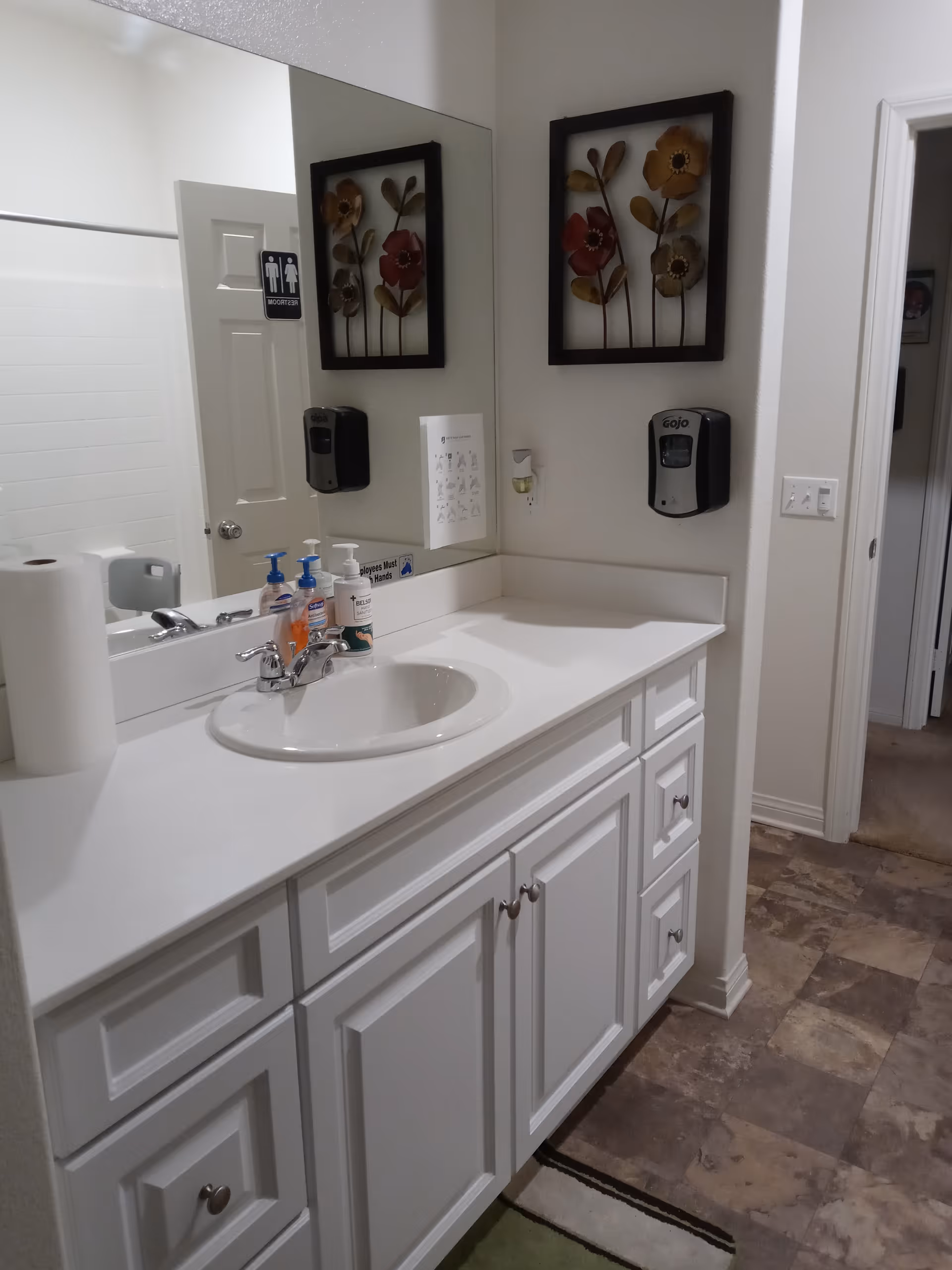A clean bathroom sink area with a white countertop and cabinetry. There are soap dispensers, a paper towel roll, and a hand sanitizer dispenser mounted on the wall. A large mirror reflects the opposite wall, which has a door labeled as a restroom. Decorative floral wall art is hung above the sink.