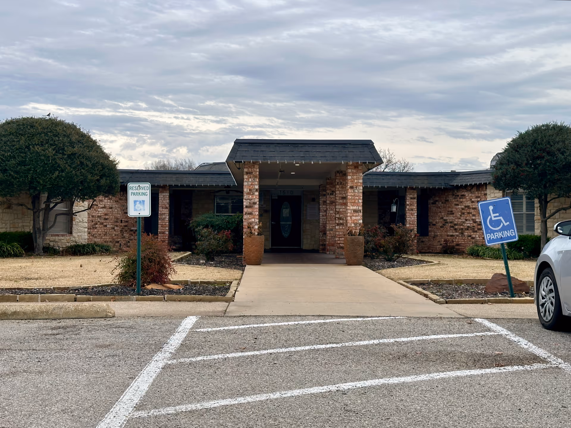 Front entrance of Lindan Park Rehabilitation Center showing a brick building with a covered walkway supported by brick pillars. There are two large potted plants near the entrance, trimmed bushes, and two reserved parking signs including one for handicapped parking. A white car is partially visible on the right side of the image.