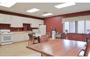 A spacious kitchen area with white cabinets, a white stove, microwave, and refrigerator. There are two wooden tables with chairs around them. The walls are painted a reddish color and there is a large window with blinds and a valance letting in natural light.
