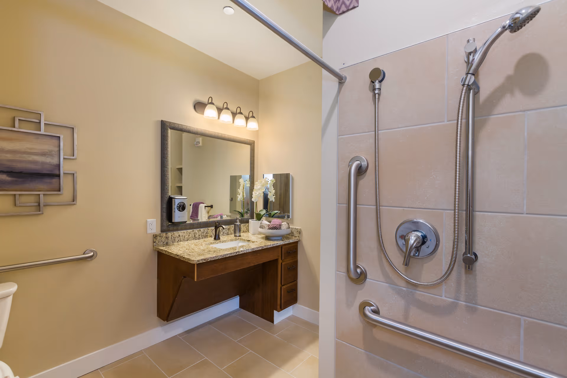 A modern bathroom with beige tiled walls and floor, featuring a walk-in shower with stainless steel grab bars and a handheld showerhead. There is a wooden vanity with a granite countertop, an under-mount sink, a large framed mirror above it, and a light fixture with four bulbs. A decorative painting is hung on the wall, and a small vase with white flowers and a bowl with towels are placed on the countertop.