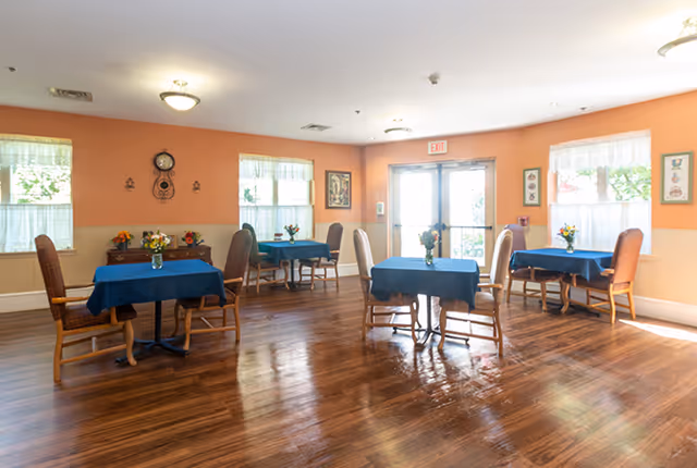 A bright dining room with wooden floors and peach-colored walls. There are four tables covered with dark blue tablecloths, each surrounded by wooden chairs with woven backs. Small flower arrangements are placed on each table. The room has several windows with white curtains allowing natural light to enter. A clock and framed pictures decorate the walls, and a glass door with an exit sign is visible in the background.
