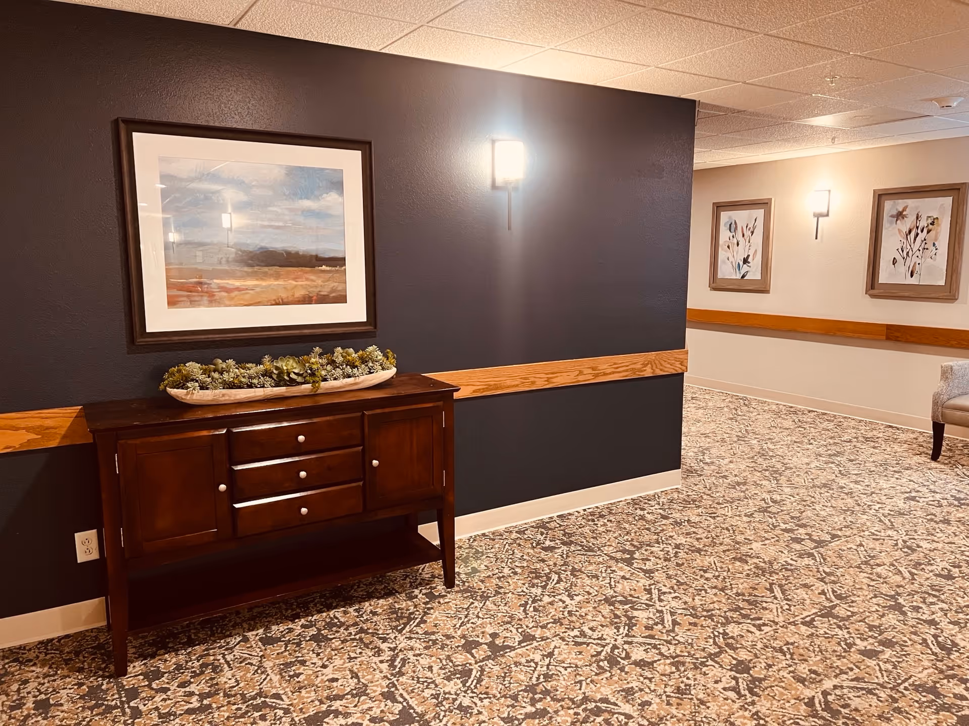 Interior hallway of a senior living facility with patterned carpet, a dark accent wall featuring a wooden sideboard with drawers and cabinets, a decorative planter with succulents on top, and a framed landscape painting above. The hallway walls are light beige with wooden chair rails and two framed floral artworks. Wall-mounted lights illuminate the space.