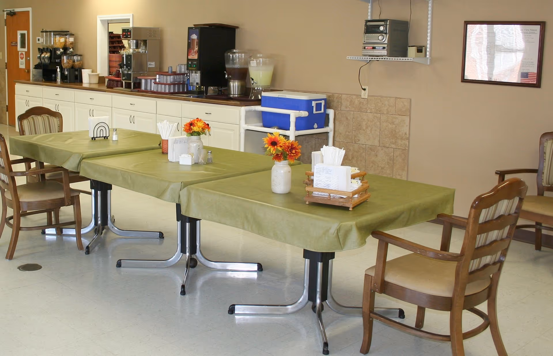Dining area with tables covered in green tablecloths, wooden chairs, floral centerpieces and a beverage station along the back counter.