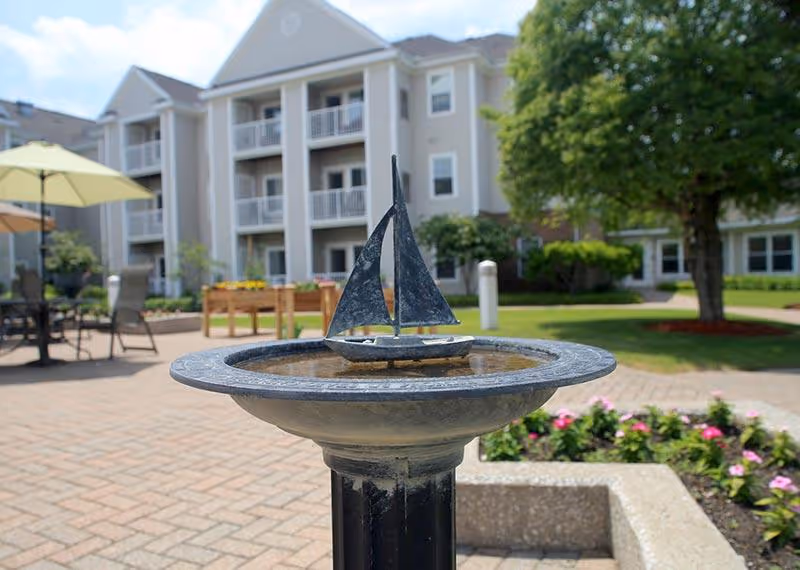 A small decorative birdbath with a metal sailboat sculpture in the center, set in an outdoor courtyard area with paved walkways, flower beds, patio furniture with umbrellas, and a multi-story residential building in the background.